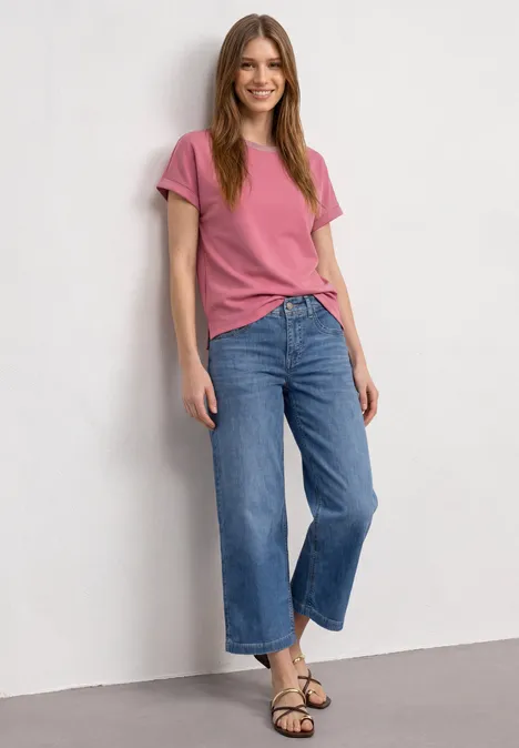 Eine junge Frau lehnt lächelnd an einer Wand, trägt ein rosafarbenes T-Shirt, weite blaue Jeans und Sandalen.