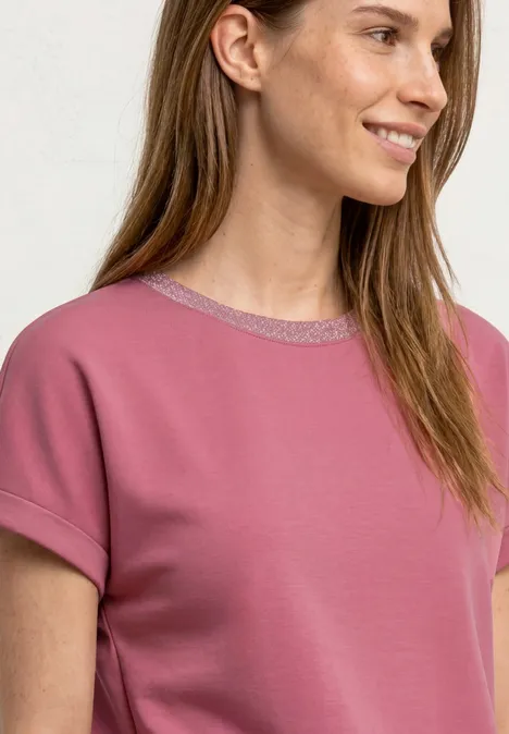 Frau in rosa T-Shirt mit kurzen Ärmeln und silbernem Kragen lächelt seitlich.