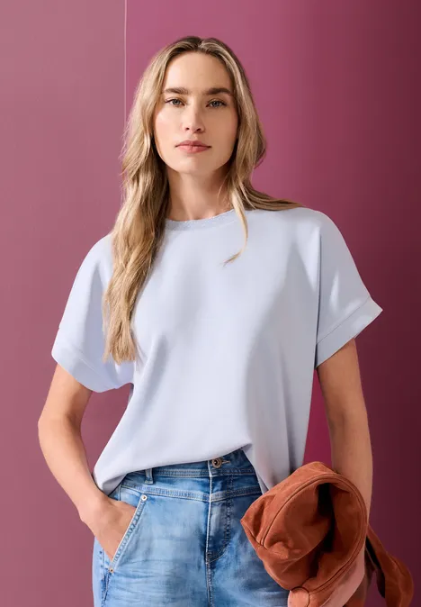 Femme en tee-shirt bleu clair tenant une veste orange devant un mur rose