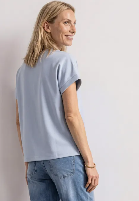 Femme souriante en T-shirt bleu et jean, debout de profil.