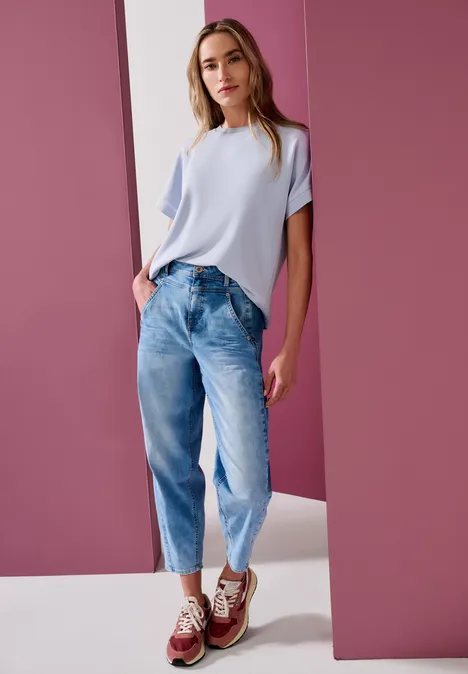 Femme portant un t-shirt clair, jean ample et baskets, posant devant un mur rose.
