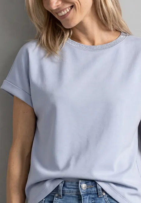 Une femme souriante porte un t-shirt bleu clair et un jean.