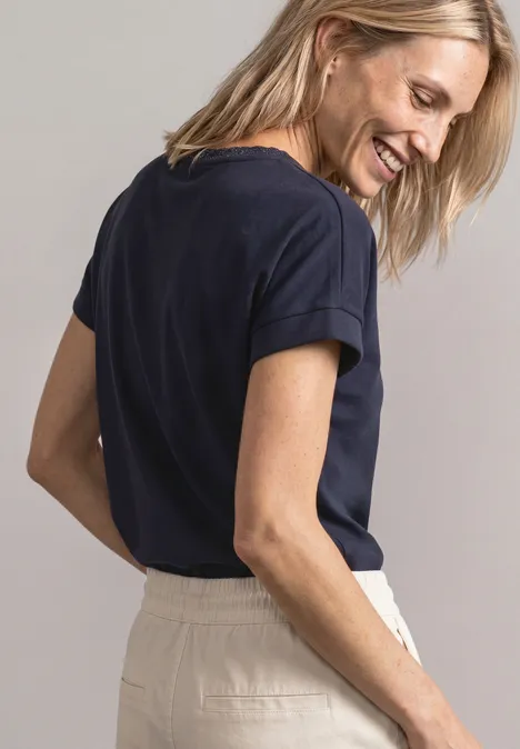 Eine lächelnde Frau in seitlicher Pose trägt ein dunkelblaues T-Shirt und helle Hose.