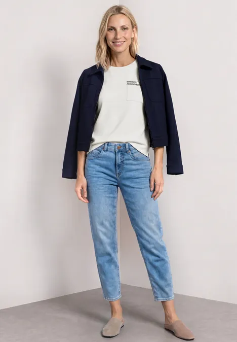 Shirt met decoratieve borstzak off white Vrouw met wit T-shirt, donkerblauw jasje op de schouders, lichtblauwe jeans en beige ballerina’s.