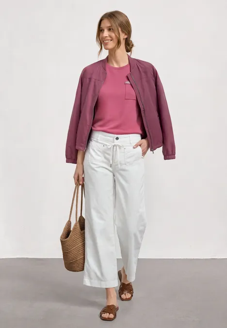 T-shirt avec poche poitrine décorative blush berry Une femme porte un t-shirt rose, une veste violette, un pantalon blanc, sandales et sac en osier en marchant.
