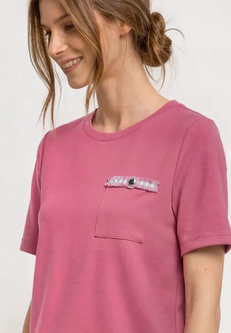 T-shirt avec poche poitrine décorative blush berry Une jeune femme souriante porte un t-shirt rose à poche poitrine orné d'un petit accessoire.