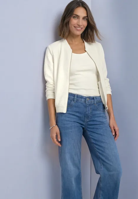Eine Frau in weißem Cardigan, Tanktop und blauen Jeans steht lächelnd vor neutralem Hintergrund.