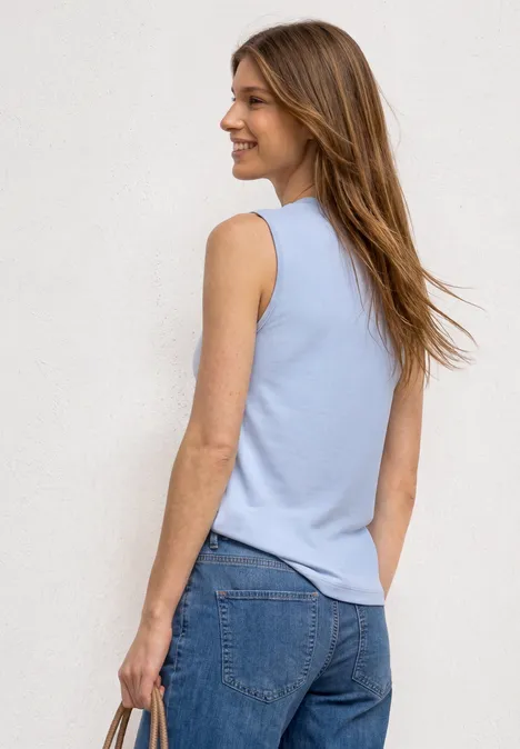 Junge Frau von hinten mit hellblauem ärmellosem Top und blauen Jeans vor weißer Wand.