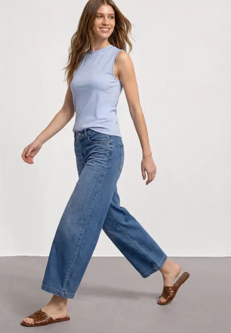 Frau in ärmellosem hellblauen Top, weiten Jeans und braunen Sandalen vor neutraler Wand.