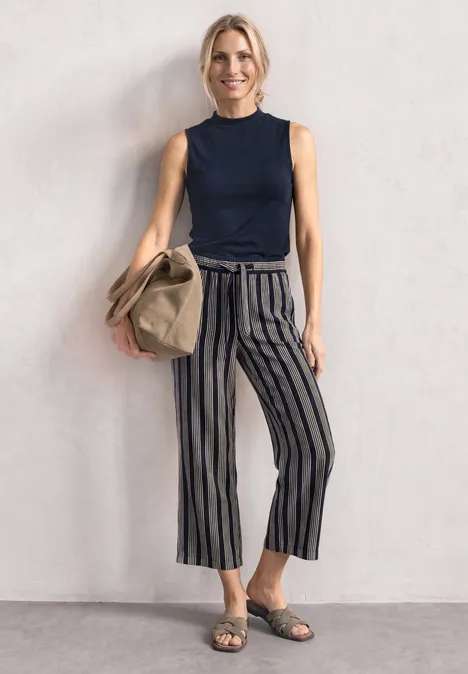 Silk-Look Top z golfem darkest blue Kobieta w granatowym topie, szerokich pasiastych spodniach, sandałach, trzyma beżową torbę.