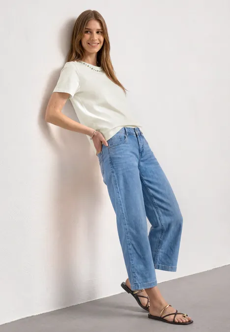 Femme appuyée sur un mur, T-shirt blanc, jean ample et sandales noires.
