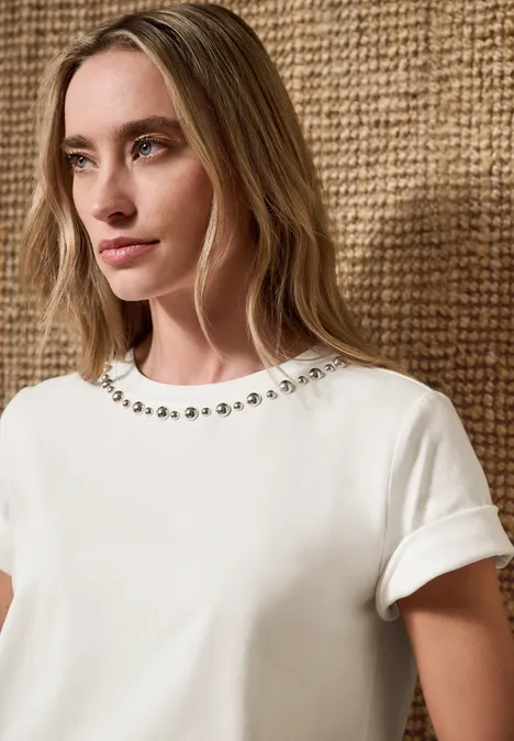 Femme en t-shirt blanc à col rond orné de perles, devant un fond tissé couleur naturelle.