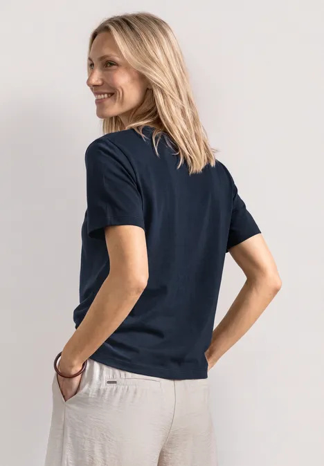 Rückansicht einer Person in dunkelblauem T-Shirt und hellbeiger Hose, Hand in Tasche.