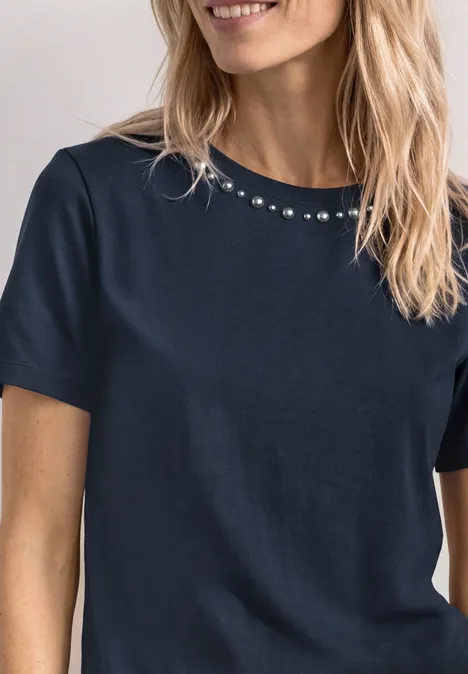Blonde Frau im dunkelblauen, kurzärmligen Shirt mit Rundhalsausschnitt und dezenten Ziersteinchen.