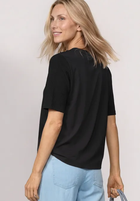 Frau in schwarzem T-Shirt und blauen Jeans, steht seitlich vor weißem Hintergrund und hält eine Tasche.