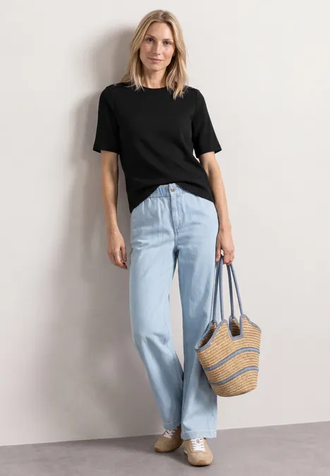 Frau mit schwarzem T-Shirt, hellblauen Jeans und Strohtasche vor weißer Wand.