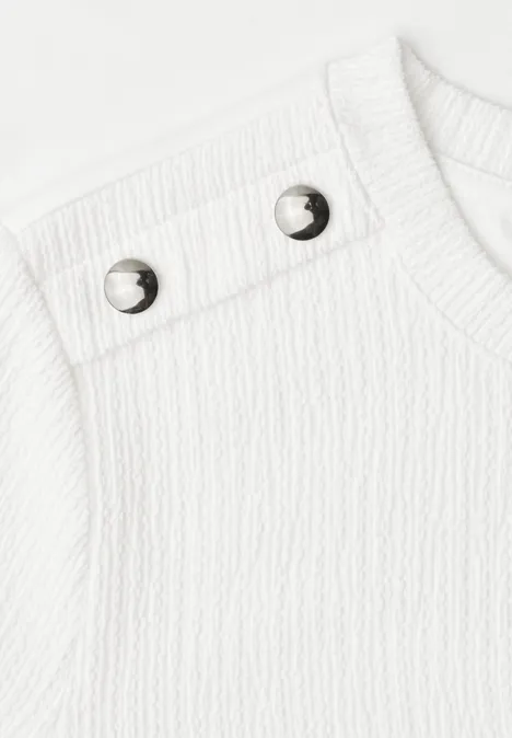 T-shirt à col rond avec boutons off white Gros plan d'un haut tricoté blanc avec deux boutons-pression argentés à l'épaule.