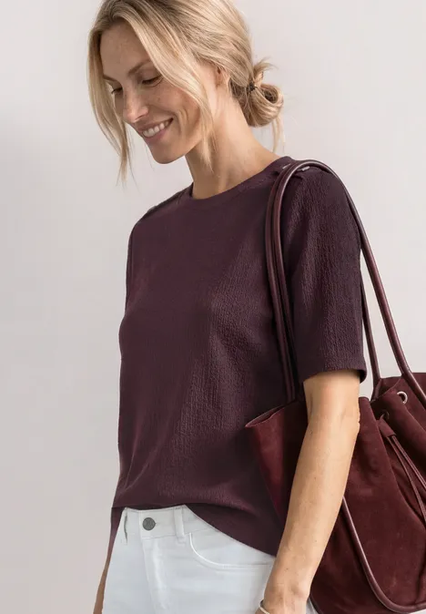 Frau in einem violetten Kurzarmshirt mit burgunderfarbener Schultertasche vor hellem Hintergrund.
