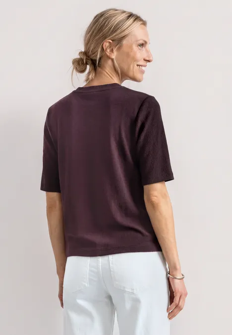 Frau von hinten in auberginefarbenem T-Shirt und weißer Hose, Haar zu einem Dutt gebunden.