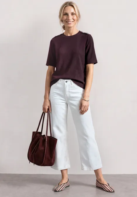 Person in kurzärmeligem dunkelrotem Shirt, weißen Cropped-Jeans, Flip-Flops und roter Handtasche.