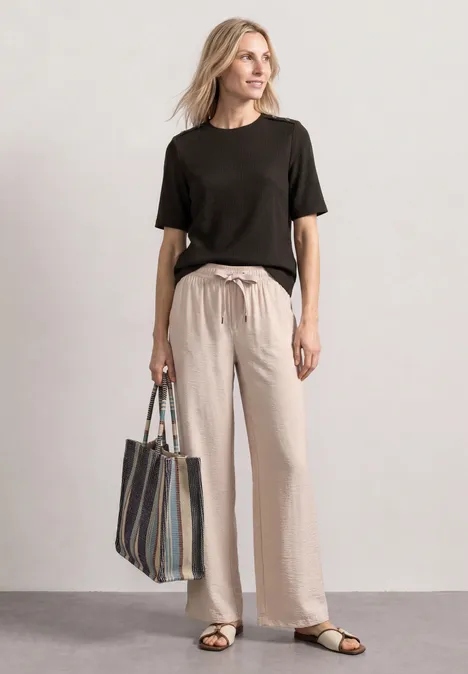 Frau in schwarzem T-Shirt und beigen Leinenhosen trägt gestreifte Tasche und Sandalen.
