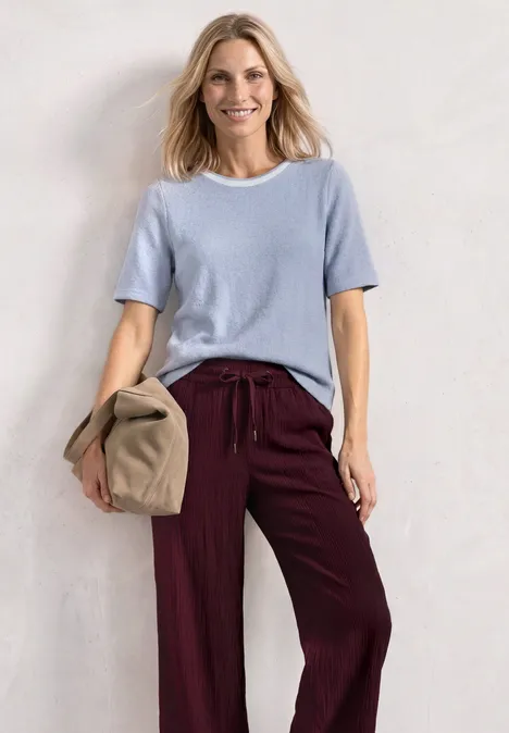 Shirt met gebreide look en ronde hals whispery blue Vrouw met lichtblauw shirt en bordeauxrode broek, houdt een beige tas vast tegen een witte achtergrond.