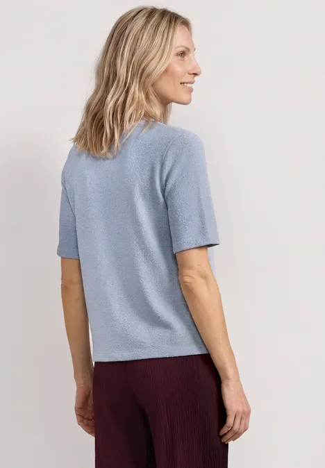 Shirt met gebreide look en ronde hals whispery blue Vrouw van achteren in lichtblauw T-shirt met korte mouwen en bordeauxrode broek, halflang blond haar.