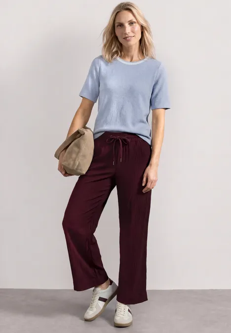 Shirt met gebreide look en ronde hals whispery blue Vrouw in lichtblauw shirt, bordeauxrode broek en witte sneakers, met een hoed in de hand.