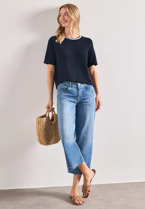 Shirt met gebreide look en ronde hals darkest blue Vrouw leunt glimlachend tegen muur, draagt zwart shirt, blauwe jeans, sandalen en een gevlochten mand.