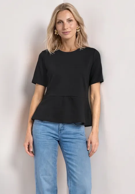Frau trägt schwarzen T-Shirt und blaue Jeans, steht frontal vor neutralem Hintergrund.