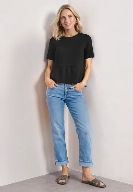 Frau in schwarzem T-Shirt, blauen Jeans und Sandalen steht vor hellem Hintergrund.