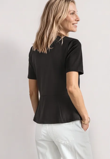 Eine Frau steht von hinten in einem schwarzen T-Shirt mit kurzen Ärmeln und weißen Hosen vor hellem Hintergrund.