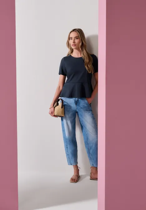 Shirt met ronde hals en volantdetail darkest blue Een vrouw draagt een zwart peplumtop, lichtblauwe jeans en sandalen, met een handtas voor een roze achtergrond.