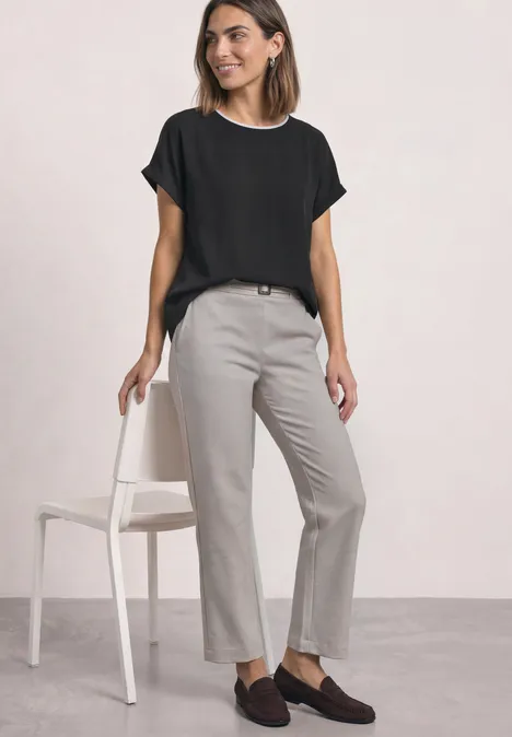 Femme en chemisier noir, pantalon gris et mocassins marron, pose près d'une chaise.