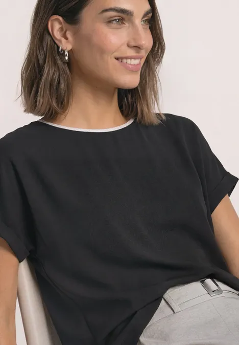 Femme en chemise noire à col blanc et pantalon gris, souriant sur le côté