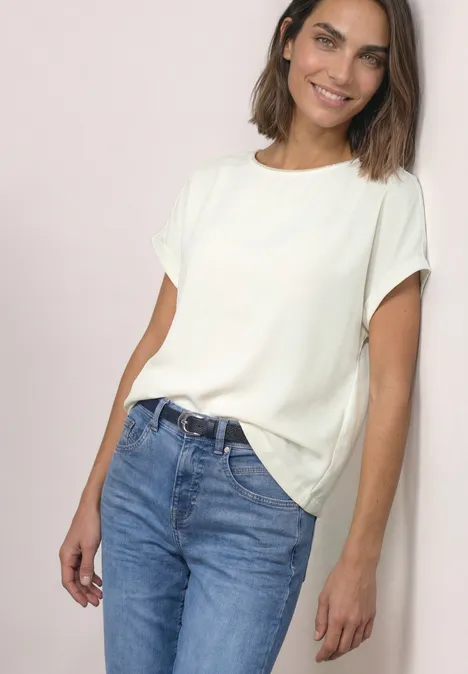 Femme devant fond clair, portant un t-shirt blanc et un jean bleu, souriante et décontractée.