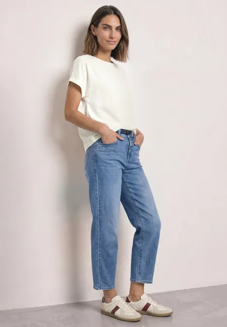 Une femme porte un t-shirt blanc, un jean bleu et des baskets blanches, posant sur fond clair.