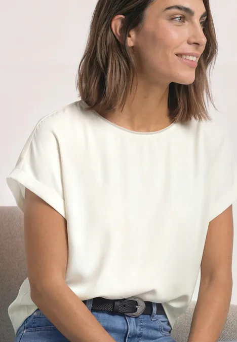 Femme souriante en t-shirt blanc à manches retroussées, jean et ceinture.