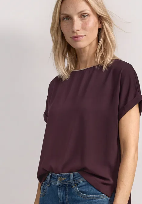Femme portant un haut bordeaux à manches courtes et un jean bleu sur fond clair.