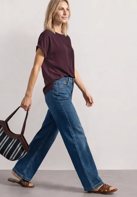 Une femme en jean et t-shirt bordeaux, portant un sac rayé, marche avec assurance.