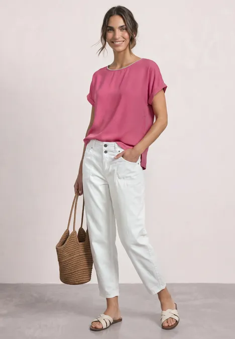 Frau trägt pinke Bluse, weiße Jeans, Basttasche und weiße Sandalen vor hellem Hintergrund.