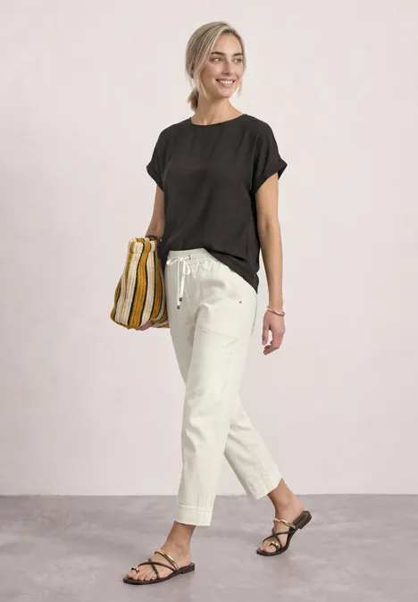 T-shirt 2 en 1 en chiffon, col bateau black coffee Femme souriante en t-shirt noir, pantalon clair à cordon et sandales, tenant un sac rayé.