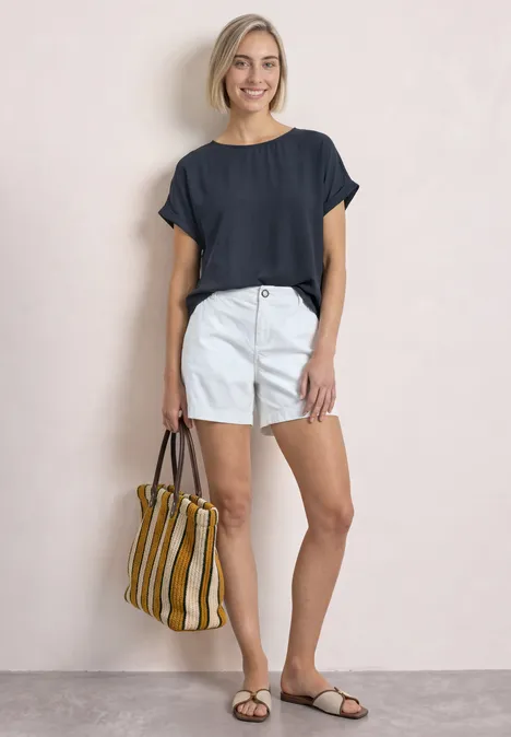2-in-1 Shirt aus Chiffon mit Boat Neck darkest blue Eine lächelnde Frau in dunkelblauem Shirt, weißen Shorts und Sandalen trägt eine gestreifte Tasche.