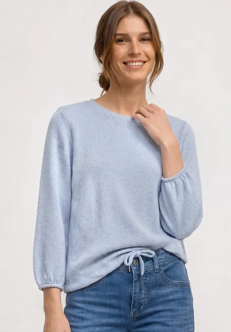 Person trägt hellblauen Strickpullover mit 3/4-Ärmeln und Bindeband, kombiniert mit blauen Jeans.