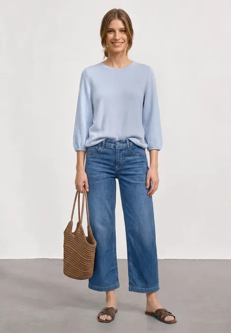 Eine Frau trägt hellblauen Pullover, weite Cropped-Jeans, Basttasche und flache Sandalen vor neutralem Hintergrund.