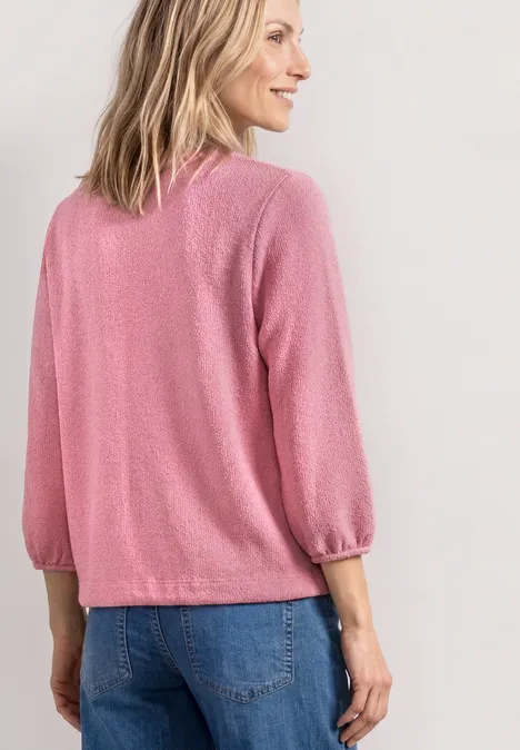 Rückansicht einer Person in rosa Pullover mit 3/4-Ärmeln und blauer Jeans vor hellem Hintergrund.