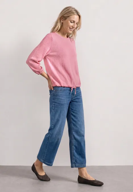 Frau in pinkem Pullover, blauen Jeans und schwarzen Ballerinas steht seitlich an einer Wand.