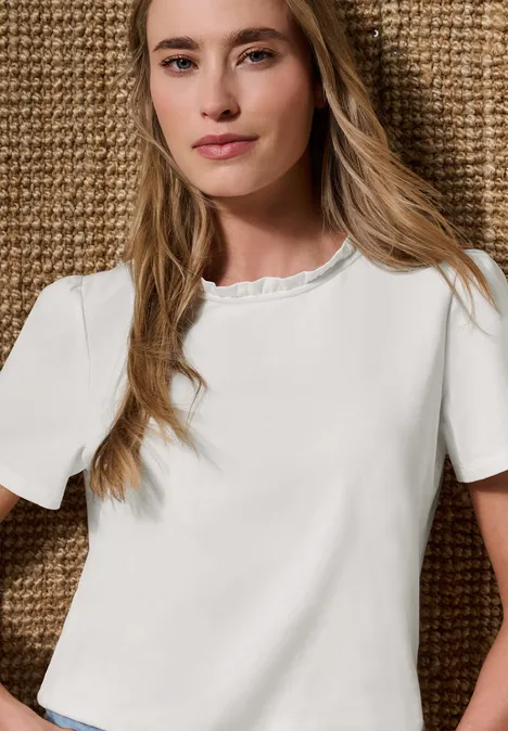 T-Shirt mit Rundhals und Rüschendetails off white Junge Frau mit langen blonden Haaren in weißem Kurzarmshirt mit Rüschenkragen vor gewebtem Hintergrund.