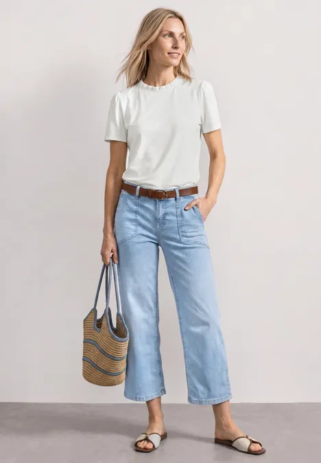 T-Shirt mit Rundhals und Rüschendetails off white Frau in weißem T-Shirt und hellblauen Jeans, trägt gestreifte Tasche und flache Sandalen vor neutralem Hintergrund.