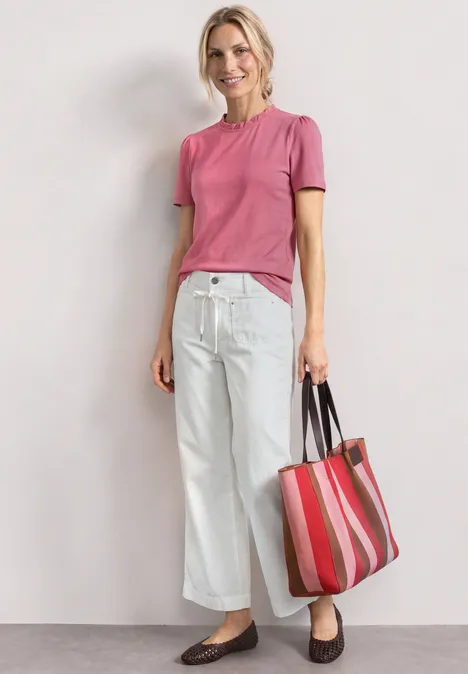 Person trägt rosa T-Shirt, weiße weite Hose, braune Slipper und hält eine rot-gestreifte Tasche.